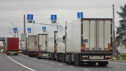 The queue traffic of many trucks with semi-trailers to the control point in the warehouse, inspector in yellow jacket check the cargo documents of the lorry drivers, weight control point