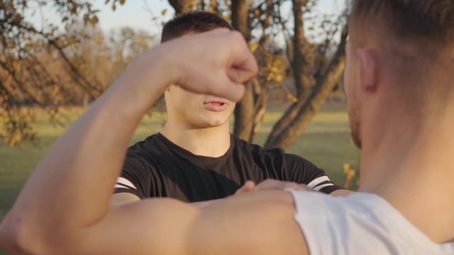 Back view of a young Caucasian sportsman boasting his muscles in sunlight in the autumn park. Young bodybuilder spending sunny day with his trainer outdoors. Healthy lifestyle concept.