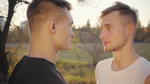 Close-up faces of two Caucasian boys looking at each other in sunlight. Adult sportsmen standing outdoors in the autumn park. Healthy lifestyle, sport concept.