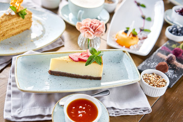 Piece of delicious cheesecake with strawberry and mint leaves on white plate. Breakfast in the cafe, morning coffee. Cappuccino and lots of desserts on the table.