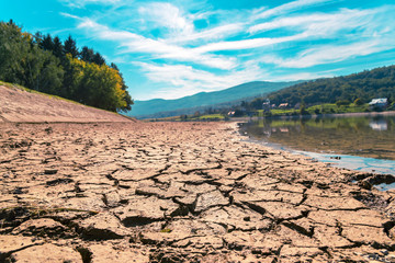 Dry cracked soil on the drying up watering place, river coast, beautiful landscape on the background - obrazy, fototapety, plakaty