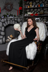 Fototapeta na wymiar A young, beautiful girl in a black dress with a skull in her hands, sits in a rocking chair near the fireplace. A model with clean skin. An attractive witch. Halloween