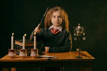 Little witch dressed in dark clothes sitting at the table against a black smoky background. There are magic wand, books with spells, candles on it.