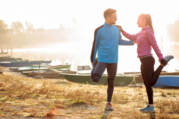 Beautiful happy young couple stretching by the lake in the early morning, warming up for jogging. Sun rising through the mist over water, rowing boats along the shore. Healthy and active lifestyle.