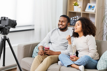 Obraz premium blogging, videoblog and people concept - happy african american couple of video bloggers with camera and smoothie drinks videoblogging at home