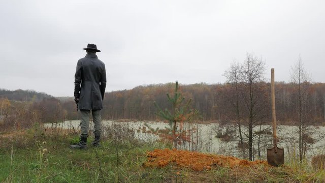 man in a black coat and hat stands near the swamp and looks into the distance