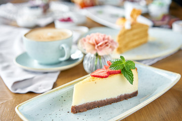 Piece of delicious cheesecake with strawberry and mint leaves on white plate. Breakfast in the cafe, morning coffee. Cappuccino and lots of desserts on the table. - obrazy, fototapety, plakaty