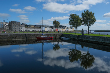 Famous view of the brightly painted houses of Galway city and the River Corrib, with sail boats moored on calm water, with the sky reflecting in it. Taken on a sunny summer day.