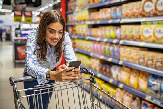 Cropped image of girl leaning on shopping cart, using a mobile phone and smiling. Woman buy products with her trolley at supermarket. Woman shopping in supermarket