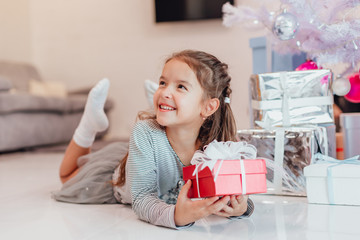 Beautiful little angel girl with perfect smile laying on the floor, holding a red present box with...