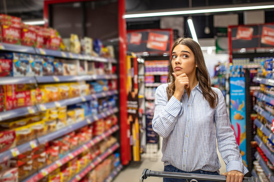 Young woman looking confused choosing products from an aisle at the supermarket walking with her trolley rubbing her chin thoughtfully copyspace people lifestyle consumerism retail buying customer