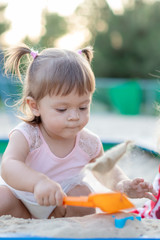 Cute toddler girl playing in sand on outdoor playground. Beautiful baby in red trousers having fun on sunny warm summer day. Child with colorful sand toys. Healthy active baby outdoors plays games