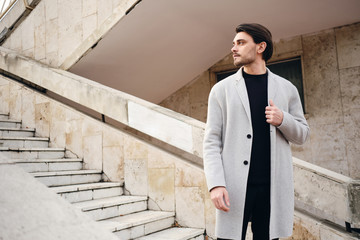 Young attractive stylish man in coat thoughtfully looking away on city street