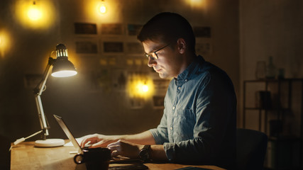 Fototapeta premium Concentrated Man Sitting at His Desk in Office Studio Working on a Laptop in the Evening. Work with Data, Analyzing Statistics.