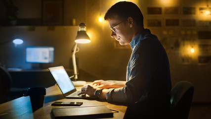 Professional Creative Man Sitting at His Desk in Office Studio Working on a Laptop in the Evening. Man working with Data and Analyzing Statistics.