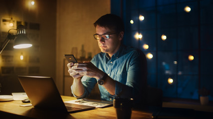 Professional Man Sitting at His Desk in Office Studio Working on a Laptop in the Evening. Man Using Smartphone with Data, Analyzing Statistics.