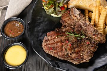 The beef of the cow grilled until cooked served with garlic bread, french frie, mixed salad and 2 dipping source. Everything is placed on a matt black plate and real wood background. Some Part of dish