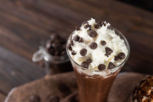 Ice chocolate, toppings by whipping cream and choc chip in a tall of clear glass on dark gray real wood and chopping board. Decorat with choc chip on floor. Popular drink to quench thirst to cool down