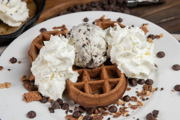 The chocolate waffle, topping with whipped cream and ice cream. Decorated with chocolate chip and cracker on white plate, on chopping real wood background. Eat sweet in moderation make to happy