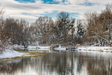 View of winter river in sunny day. Snow covered tall trees on the riverside against blue sky with white clouds