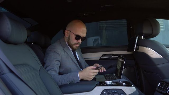 handsome businessman using his mobile phone in a modern car with a driver in center of the city. Concept of business, success, traveling, luxury 