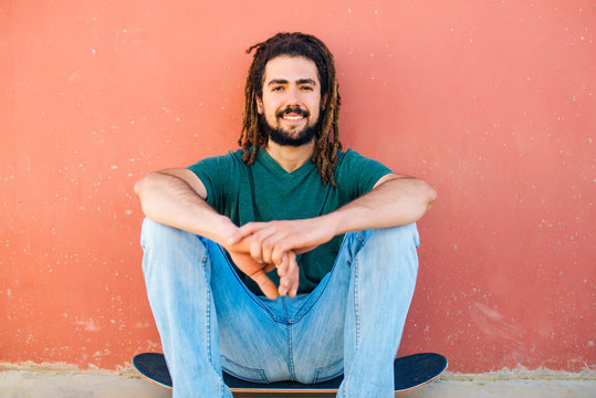Portrait of smiling young man with dreadlocks and beard sitting on his skateboard in front of a reddish wall