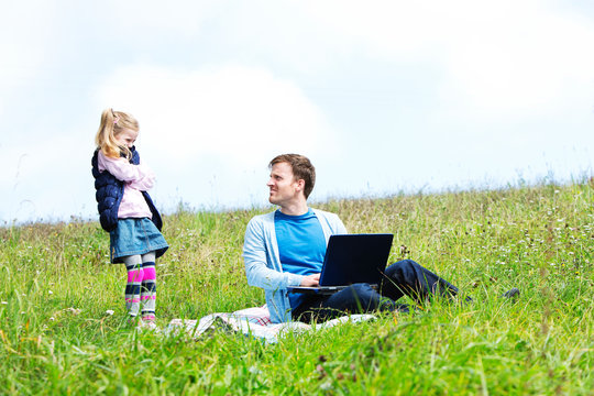 Father and daughter sitting on meadow, laptop, father looking to daughter