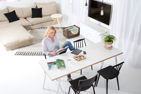 Smiling woman sitting at the table in her living room reading magazine