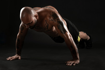 Obraz premium Athletic bald, tattooed man in black shorts and sneakers is posing against a black background. Close-up portrait.