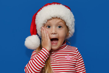 Portrait of surprised and shocked little toddler child girl in Santa hat standing isolated over blue background. Looking at camera. hands near open mouth. Happy New Year and Christmas holiday concept	