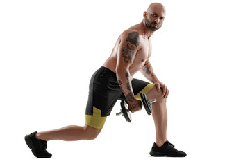 Athletic bald, tattooed man in black shorts and sneakers is posing with a dumbbell isolated on white background. Full length portrait.