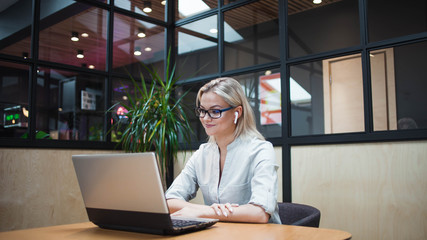 Plakat young blonde woman sits at her laptop and uses wireless headphones.