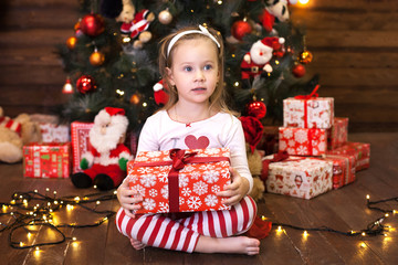 Merry Christmas and happy holidays. New Year 2020. Christmas holiday concept. Cute little girl in red striped pajamas sitting by Christmas tree with a gift. Happy kid enjoys holiday. Winter holidays. 