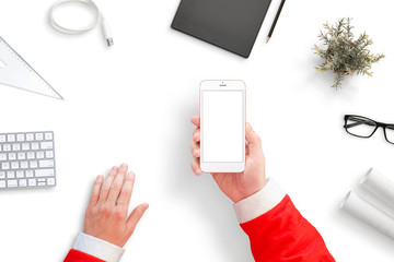Santa holding smart phone with isolated screen for mockup. Santa's office desk with papers, keyboard, folder, charger cable... Top view, flat lay.