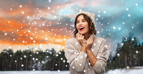 christmas, season and people concept - happy smiling young woman in knitted hat and sweater over winter forest and snow background