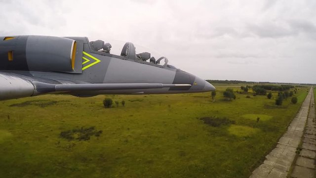 A gray blue training fighter aircraft fly quickly and low above the ground against a dark cloudy sky. Dangerous trick over the runway. View from the right wing close up.