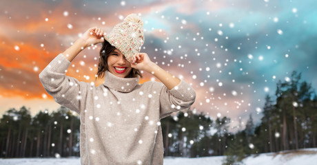 christmas, season and people concept - happy smiling young woman in knitted hat and sweater over winter forest and snow background