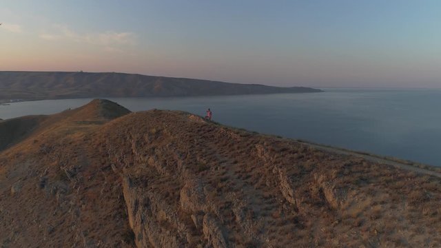 Aerial fly around girl enjoys mountain view standing on the top of the mountain. During sunset. Sea on abackground