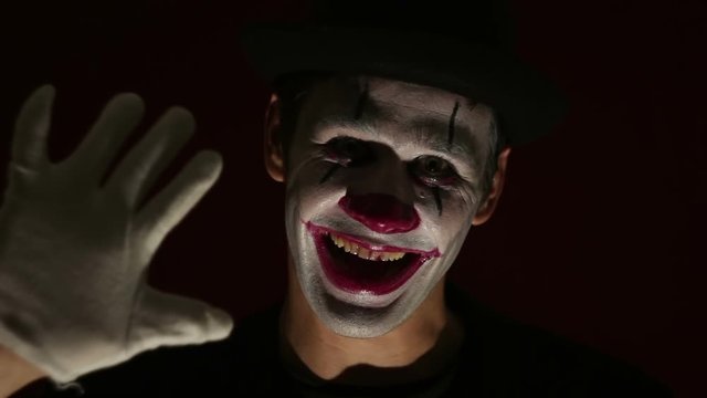 Terrible man in a clown makeup takes off his hat by welcoming his victim, looks at the camera and laughs.Terrible clown looks at the camera and laughs terribly. Scary clown grimaces looking into camer