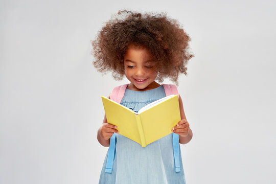 childhood, school and education concept - happy little african american girl with backpack reading book over grey background