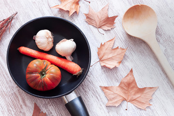 PAN WITH CARROT, GARLIC AND TOMATO NEXT TO WOOD SPOON AND TREE LEAVES ON CLEAR WOODEN TABLE. AUTUMN KITCHEN