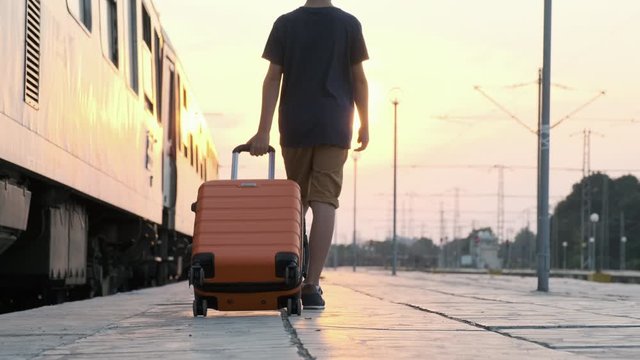 Slow motion happy boy walks along the platform of the train station and rolls an orange suitcase behind him in the rays of a bright sunny sunset. The child goes vacation by train. Tourism, travel