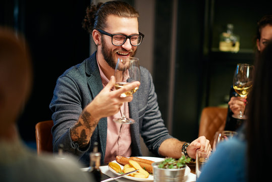 Handsome Caucasian Bearded Hipster Man With Eyeglasses Smiling And Holding Glass Of Wine While Sitting With His Friends In Restaurant.