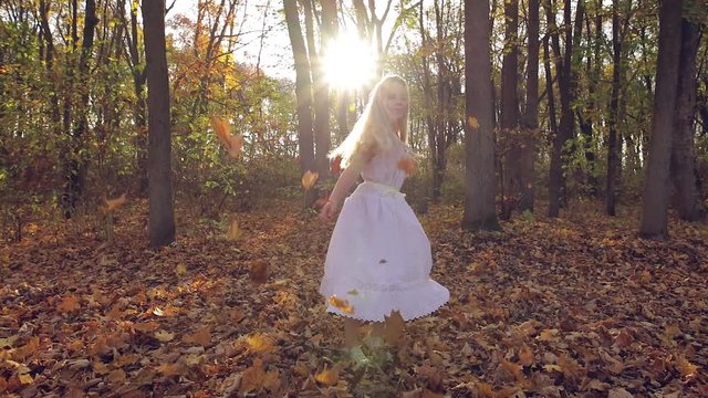 little blonde girl in white dress throws fallen leaves from trees slow motion