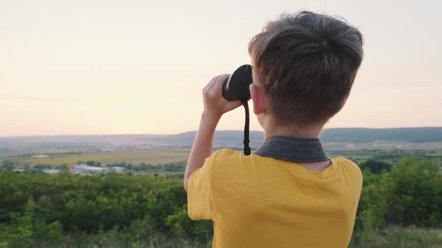 Happy boy looks through a binoculars a beautiful landscape of green hills at sunset on a sunny day in summer and joyfully says something and shows his hand on a take-off plane an airport