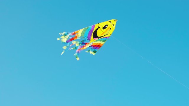 A colored kite with a smiley emoji hangs in the air against a blue sky on a sunny summer day at a city kite festival. The tail of a kite sways in the wind. Children's holiday. Lifestyle
