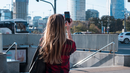 Fototapeta na wymiar Tourism concept. Young woman with beautiful hair taking photo on smartphone while walking by the city.