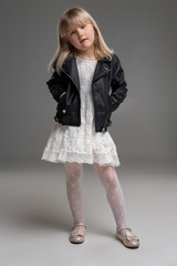 Little blonde girl in a white dress and black leather jacket is posing standing over a gray background.