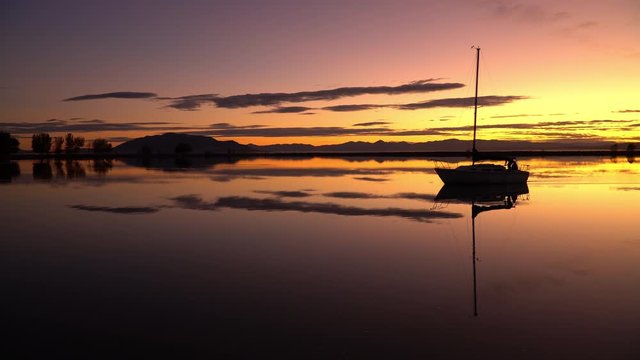 Sailboat reflection at sunset with vibrant colors in Utah Lake as it moves over the glassy water in silhouette.