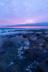 Water flowing between rocks in the coastline with cloudy sunrise sky.
